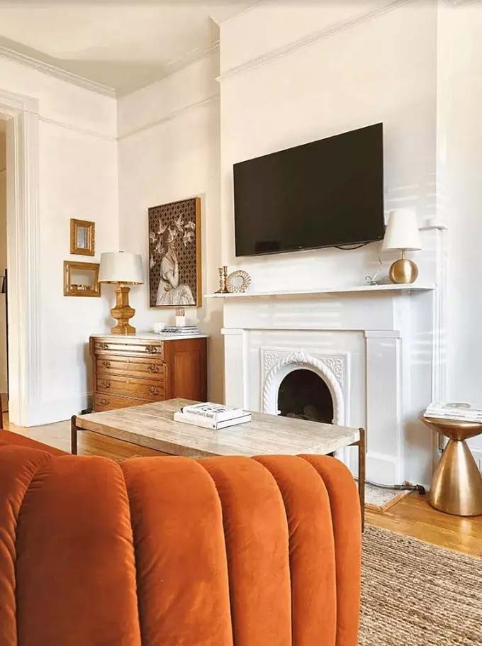 Placing the screen above a classic white fireplace saves a lot of floor space in a small room. A small lamp and leaning art pieces make the mantel look very pretty and lived-in. The bright orange sofa adds a fun pop of color to the white surroundings.