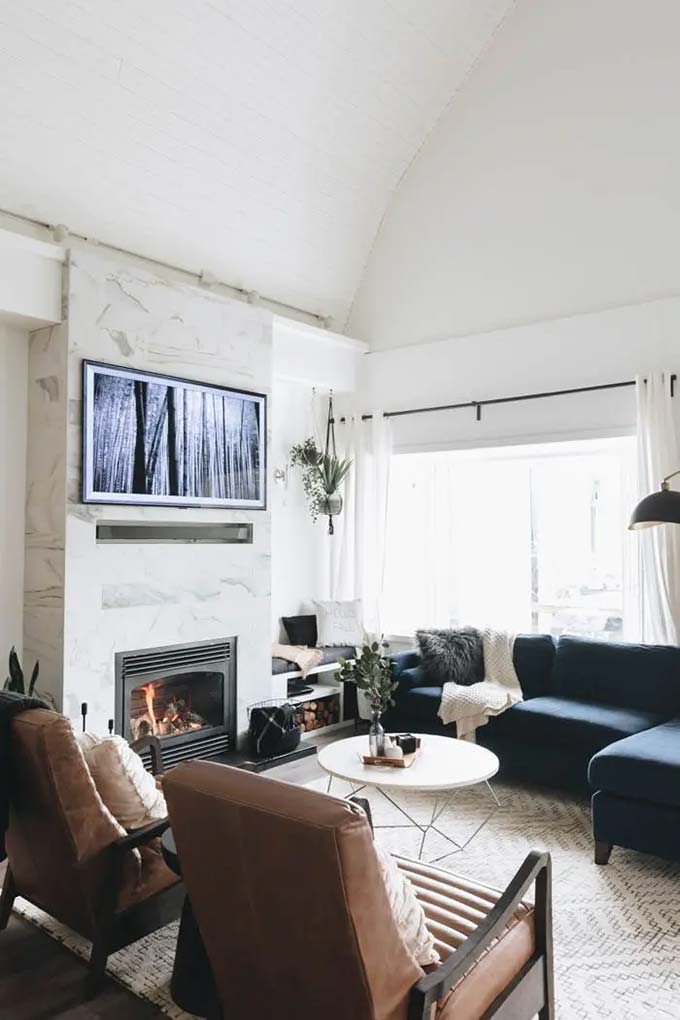 A tall wall of white marble surrounds the fireplace and the mounted screen. The dark blue sofa looks very cozy in this big, bright room with high vaulted ceilings. Using stone is a very fancy way to design a modern TV wall.