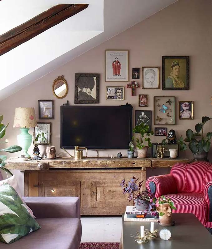 An old, heavy wooden workbench serves as a unique stand for the television under the slanted ceiling. The wall is covered in quirky art pieces, mirrors, and butterfly frames. This eclectic TV wall decor turns an awkward attic space into a cool and artistic hangout spot.