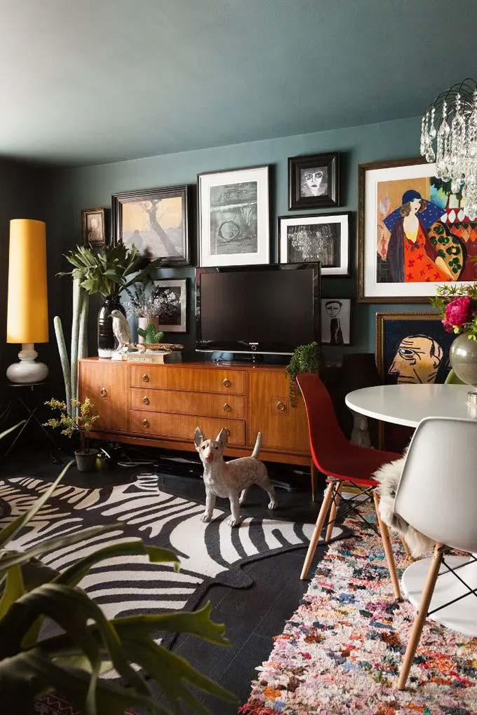 Dark teal paint makes the wooden furniture and colorful art pop beautifully. A mix of unique paintings surrounds the television to hide it within a busy gallery wall. The zebra rug and dog statue add a lot of personality to this eclectic TV wall decor.