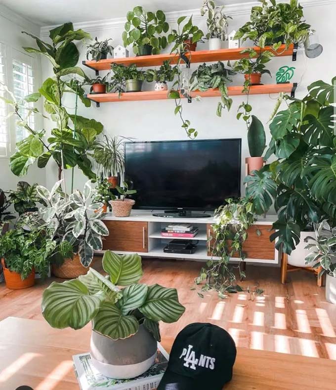 Two long macrame hangings frame the television perfectly on the white wall. A floating wooden shelf keeps the floor clear and makes the room feel much bigger. This simple TV wall decor style looks very calm with the leather poufs and olive tree.