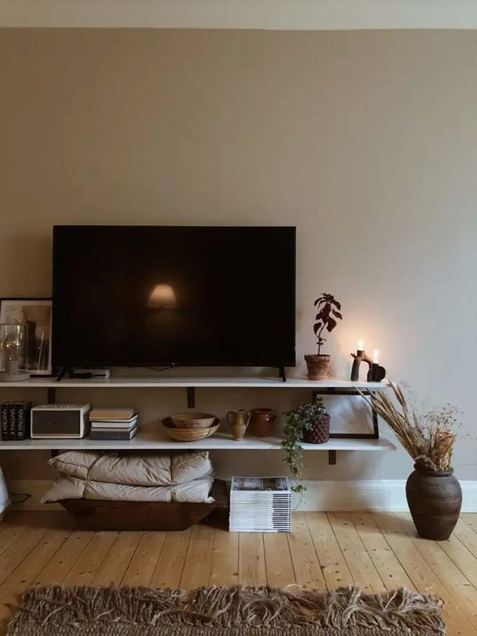 The dark gray wall makes the TV screen almost disappear into the background. Square shelf boxes arranged on the left side hold cute cactus plants and pictures. This moody TV wall decor uses industrial styles and rustic wood for a cool look.