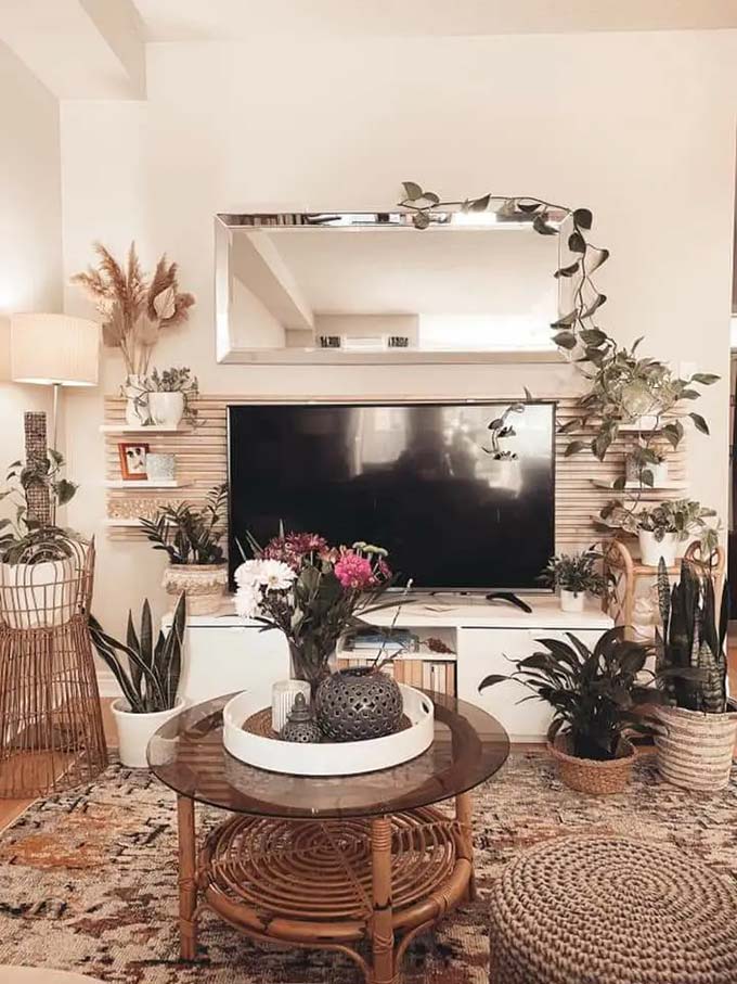 Wood slats behind the TV add texture without needing to paint the wall. A long rectangular mirror hangs above to reflect light and make the room feel bigger. Rattan furniture and plenty of plants complete this relaxing boho TV wall decor.