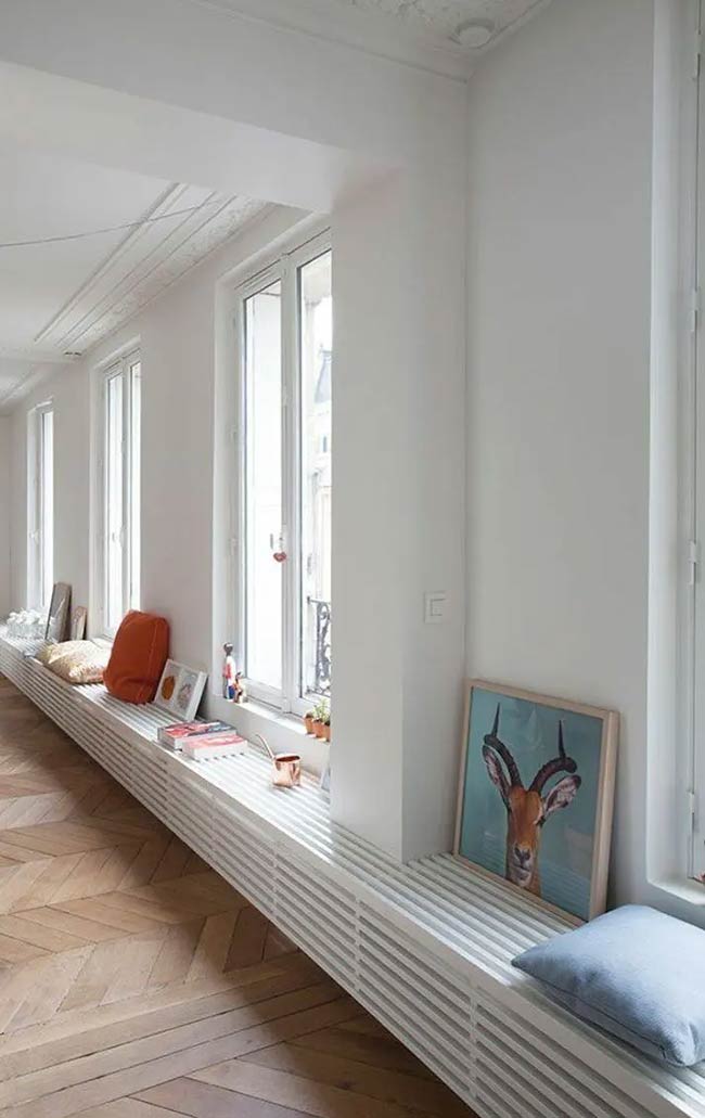 This super long bench covers the radiator and runs across two tall windows to create a seamless look. It is a simple way to add seating without blocking the beautiful view outside. The mix of colorful pillows and leaning art makes the white slats look very cozy.
