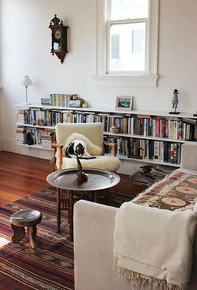 A long shelf runs under the window to hold a ton of colorful books. It fits perfectly behind the vintage armchair and keeps the white wall from looking empty. This is a smart under window storage solution for small spaces that need organization.