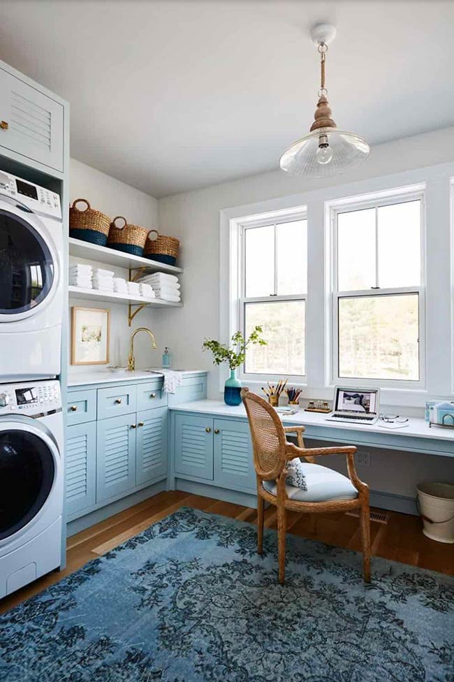This renovated laundry room doubles as a small office. Pale blue cabinets and natural wood flooring connect the space visually, while wicker chairs and baskets add warmth. A large window brings in natural light, making the room feel open, bright, and productive for both chores and work.