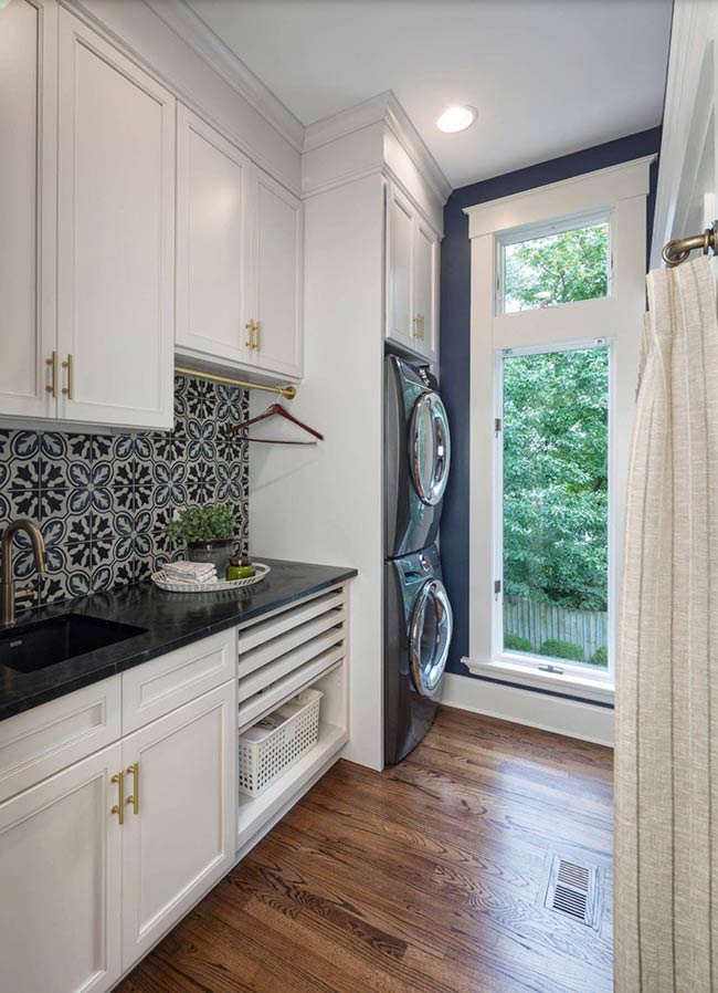 This small yet stylish laundry room proves that one bold feature can transform the space. A navy backsplash paired with black soapstone countertops gives energy and sophistication. Five custom drying racks and pull-out laundry baskets make air-drying and sorting easy. Warm red oak floors add a natural touch, balancing the cool tones above. Even in 8 by 6 feet, smart design choices create a room that is functional, organized, and visually striking, showing that size doesn’t limit style.